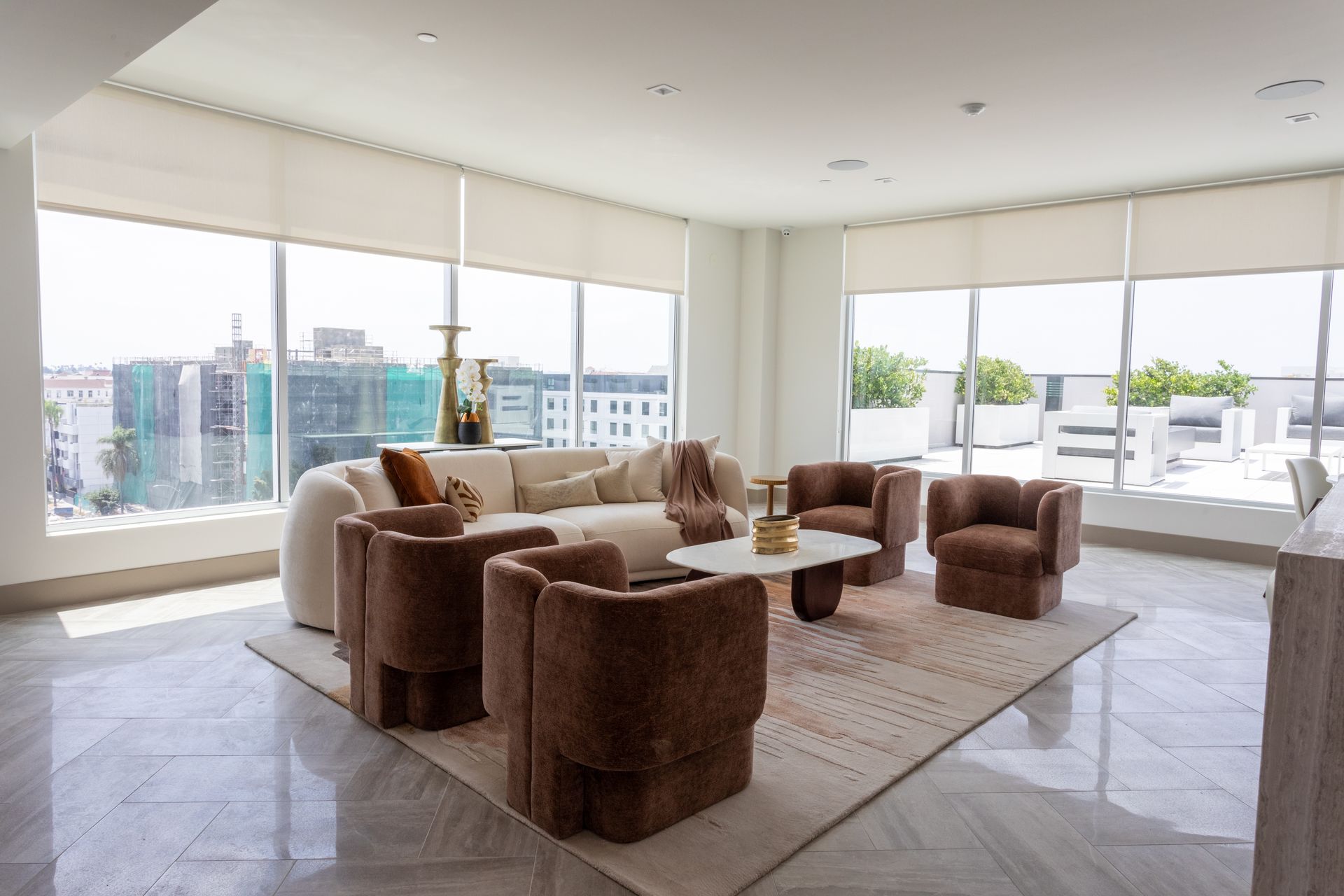 Spacious living room with large windows, light-colored sofa, and brown armchairs; a rug and coffee table are in the center.