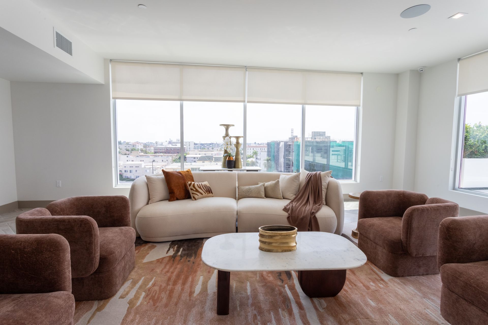 A modern living room with a cream sofa, brown chairs, and a white coffee table in front of large windows.