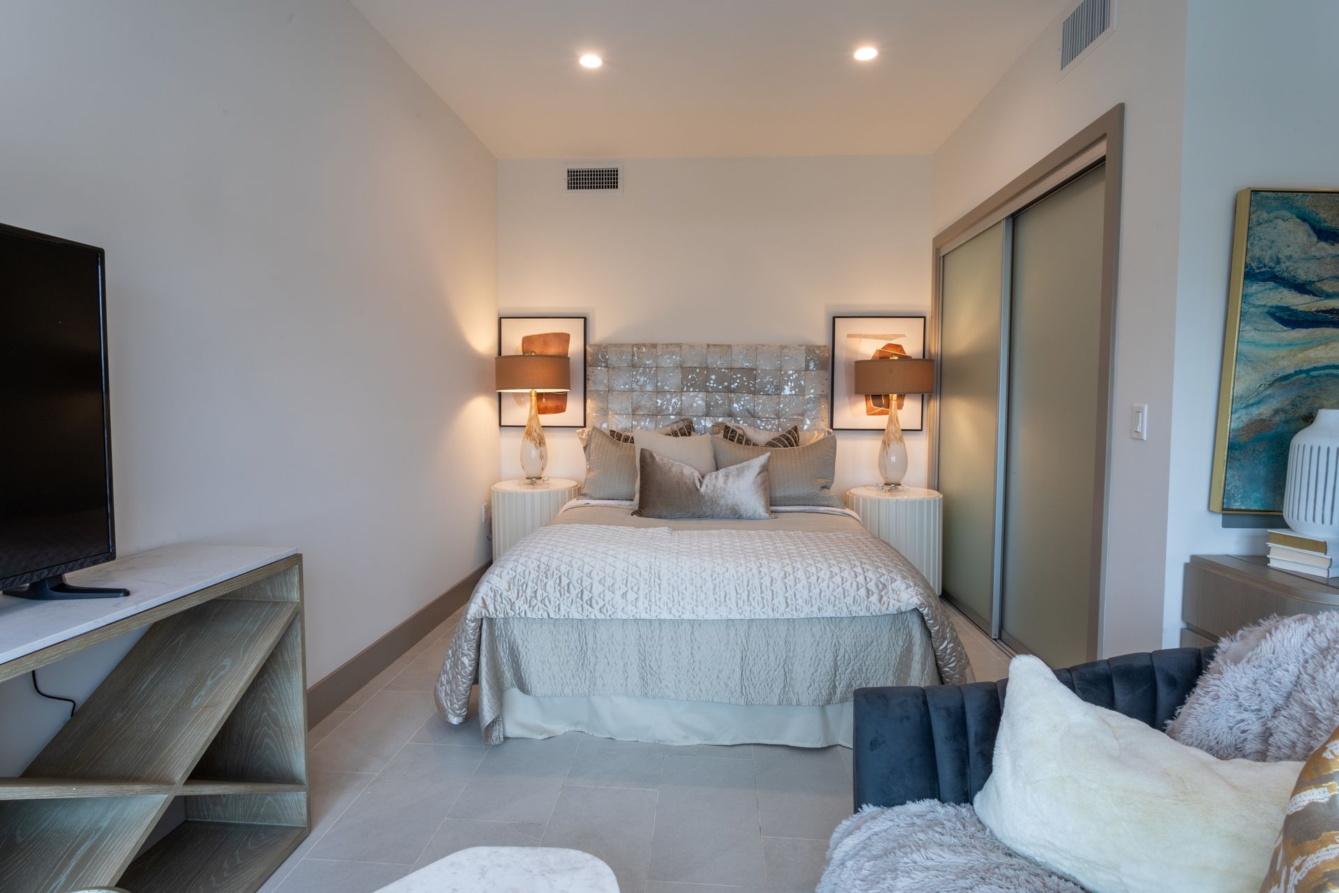 Bedroom with bed, nightstands, TV, couch, artwork, and sliding door. Neutral color palette.