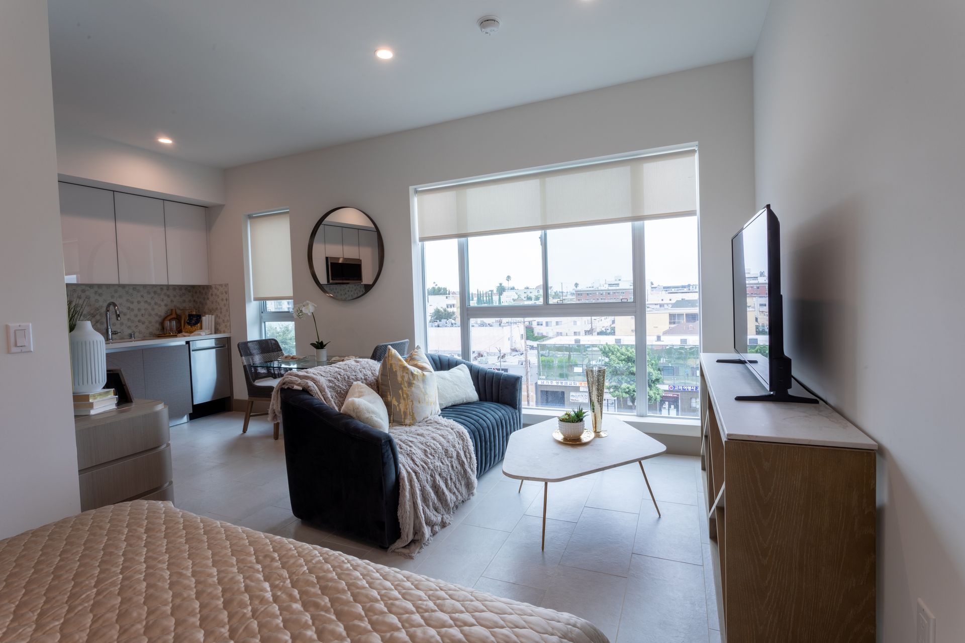Living room with sofa, TV, coffee table, and kitchen in the background. Large window.
