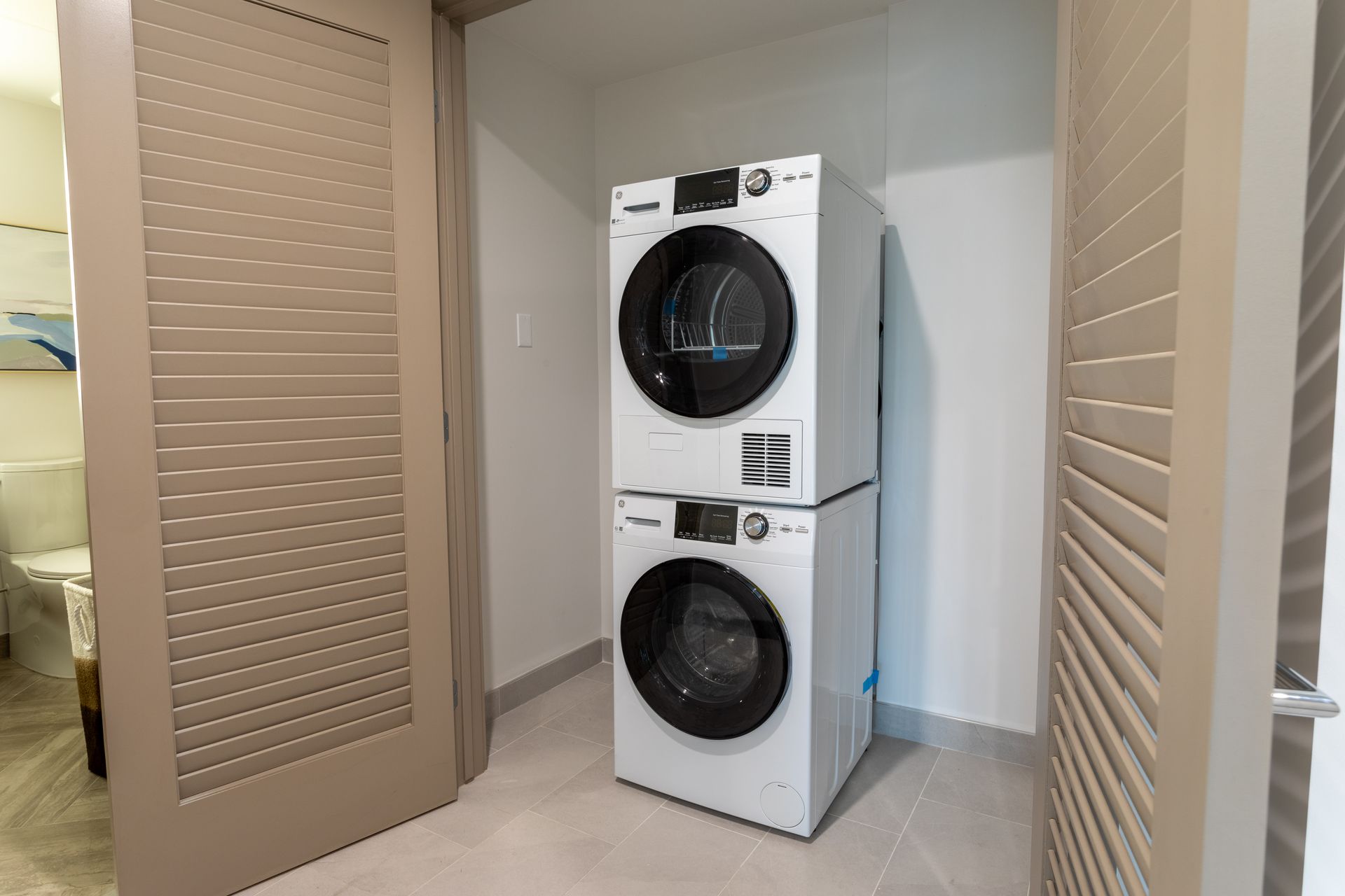 Laundry room with stacked white washing machine and dryer, nestled between two louvered doors.