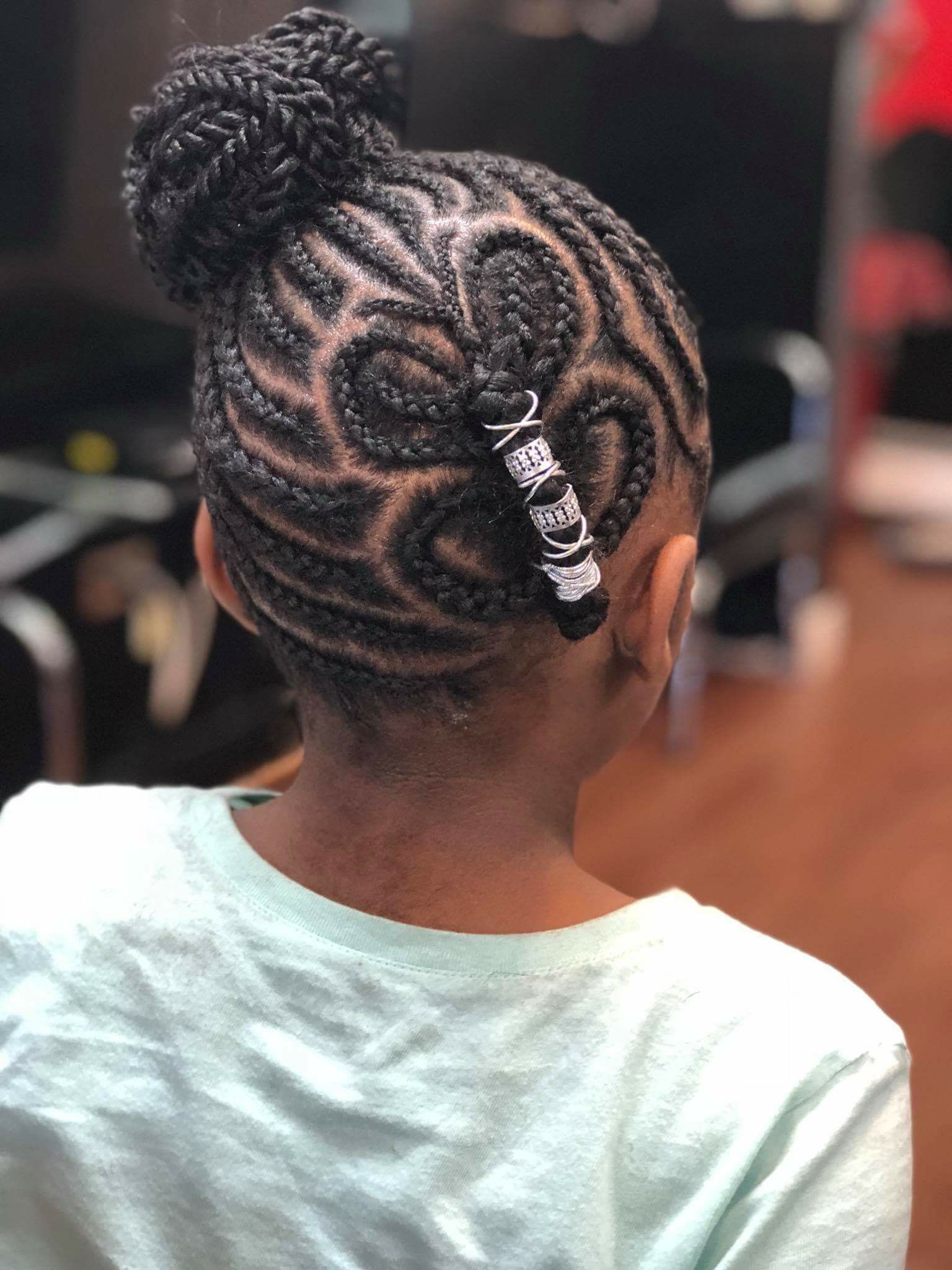Girl with intricately braided hair in a bun. Braids include swirl designs. She wears a light green shirt.