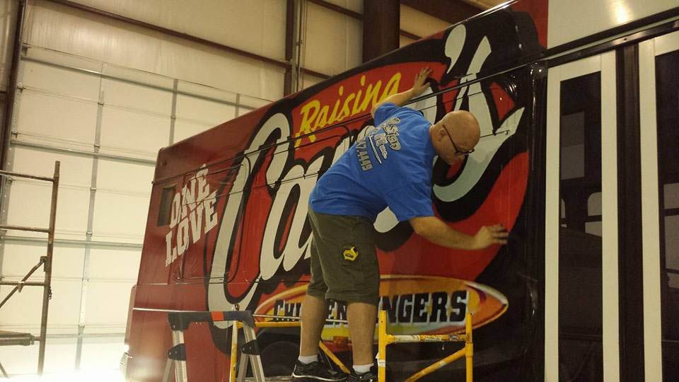 A man in a blue shirt is standing on a ladder painting a sign on the side of a truck.