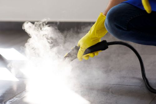 A person wearing yellow gloves is cleaning the floor with a steam cleaner.
