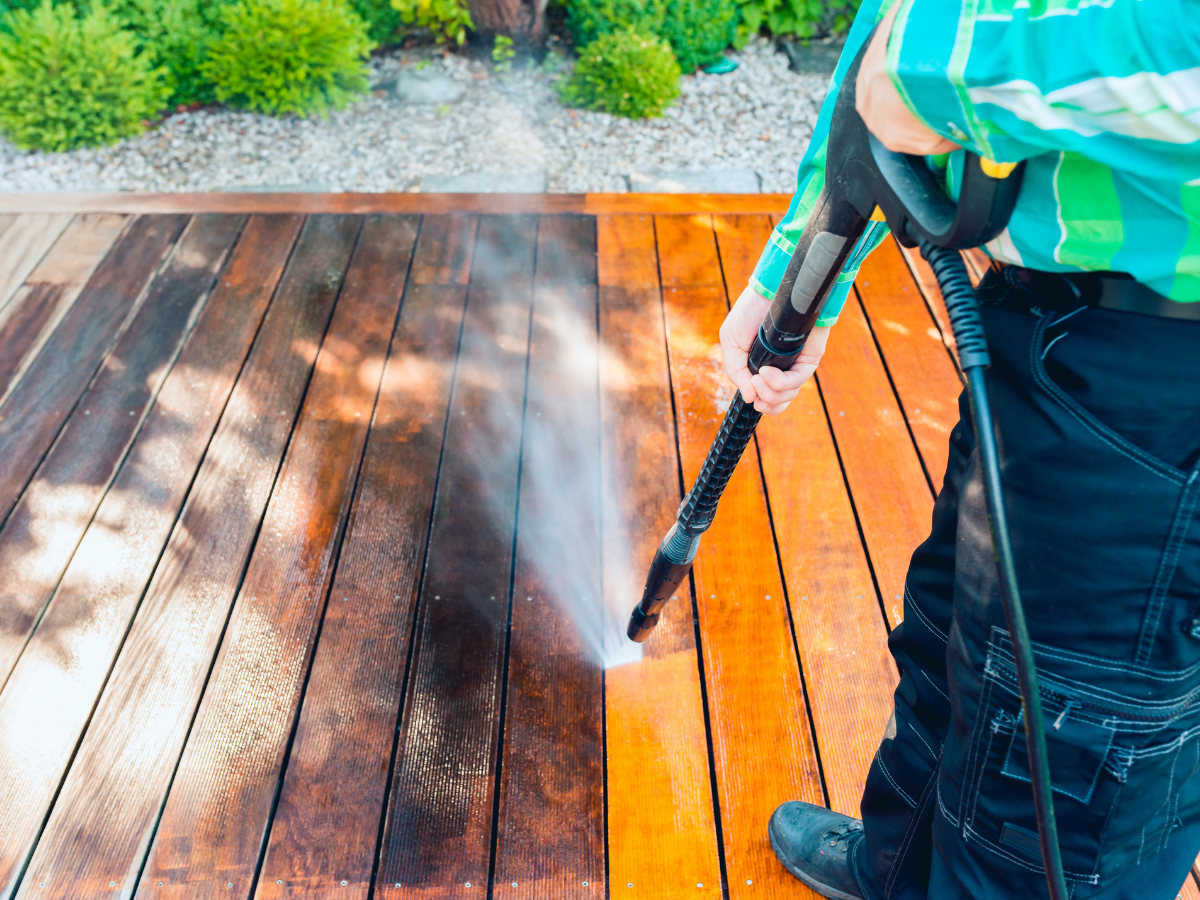 Person power washing a wooden deck, cleaning the dark side to reveal a light, clean side.