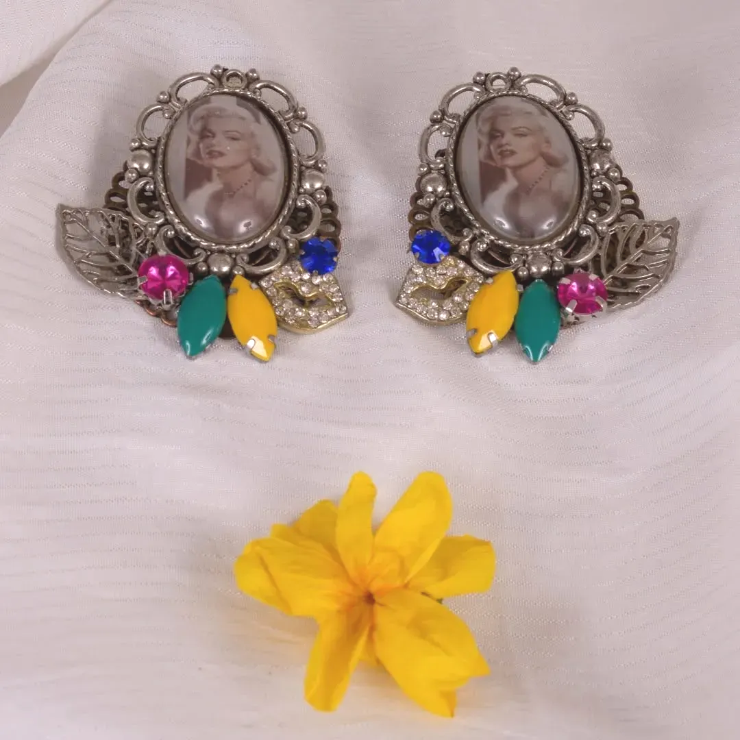 Par de pendientes ornamentados con retratos de Marilyn Monroe, gemas de colores y una flor amarilla sobre tela blanca.