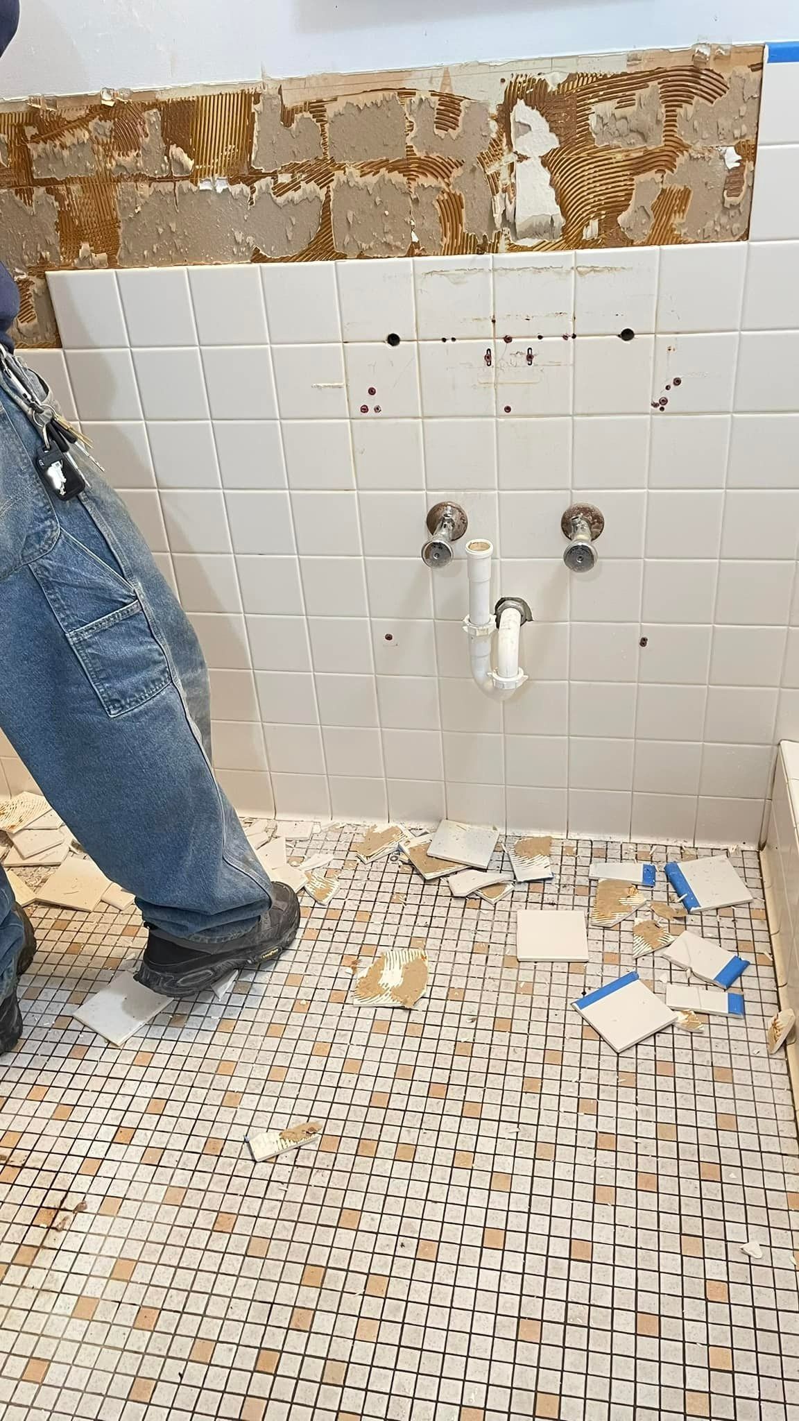 Person in jeans removes tiles from a bathroom wall. Debris covers the floor with exposed plumbing.