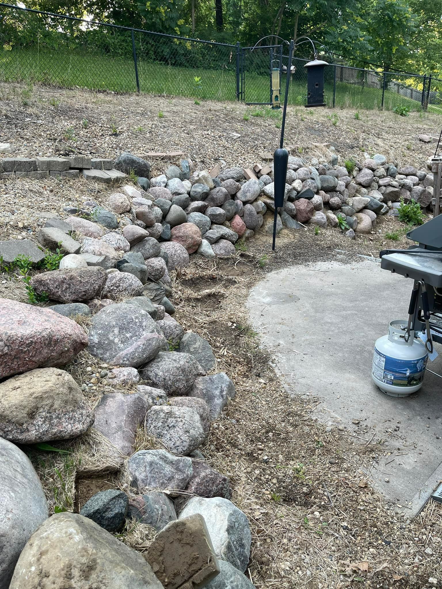 Rocks and gravel border a concrete patio and path. A grill and propane tank are visible.