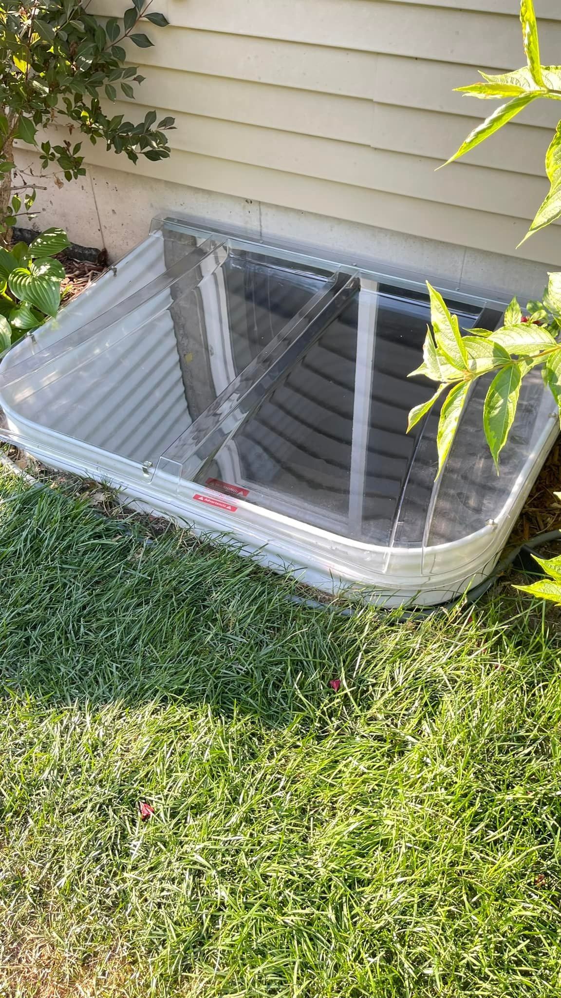 Metal window well with mesh cover next to beige siding and grass.