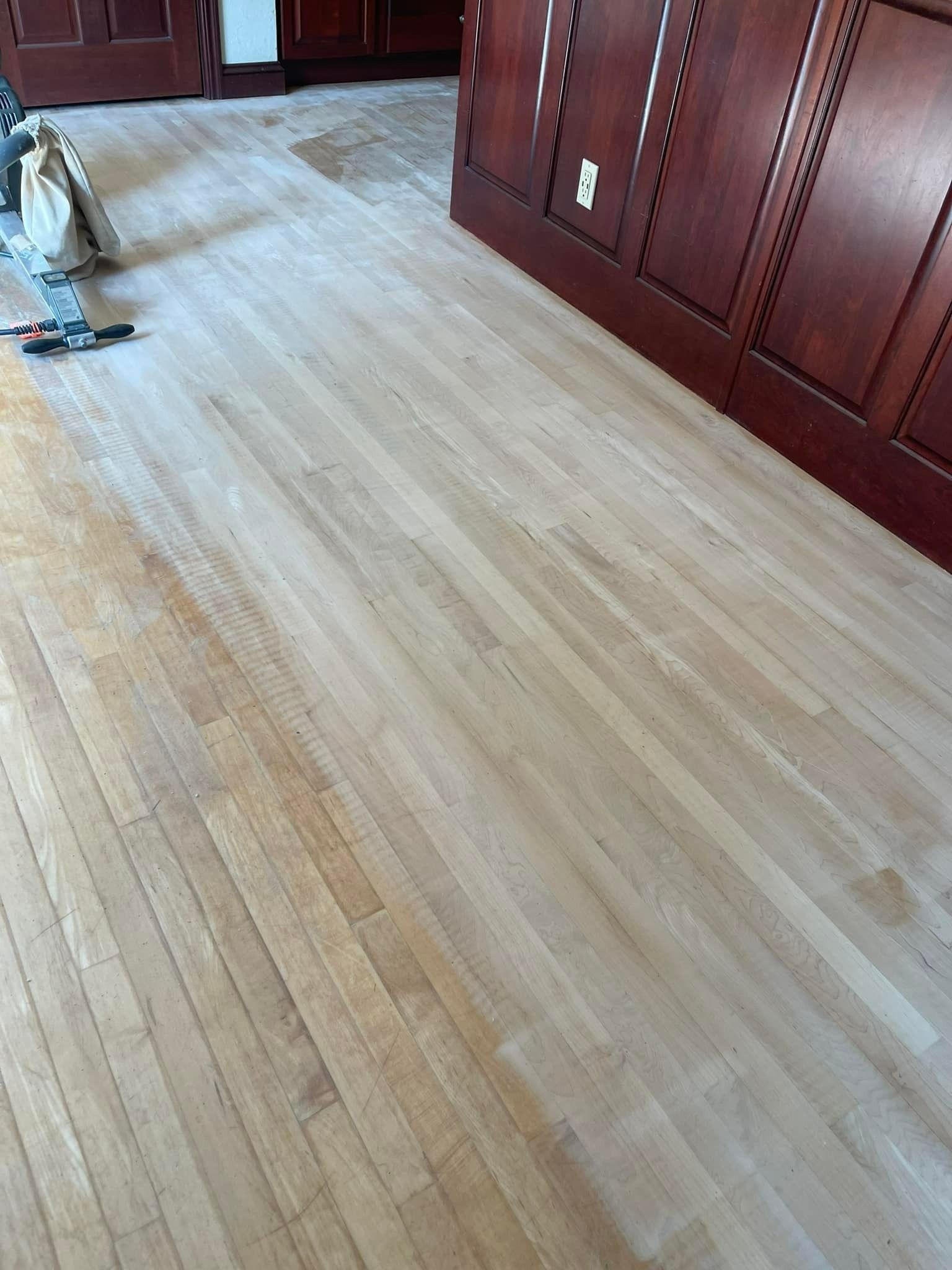Wooden floorboards being sanded, near a dark wood cabinet.
