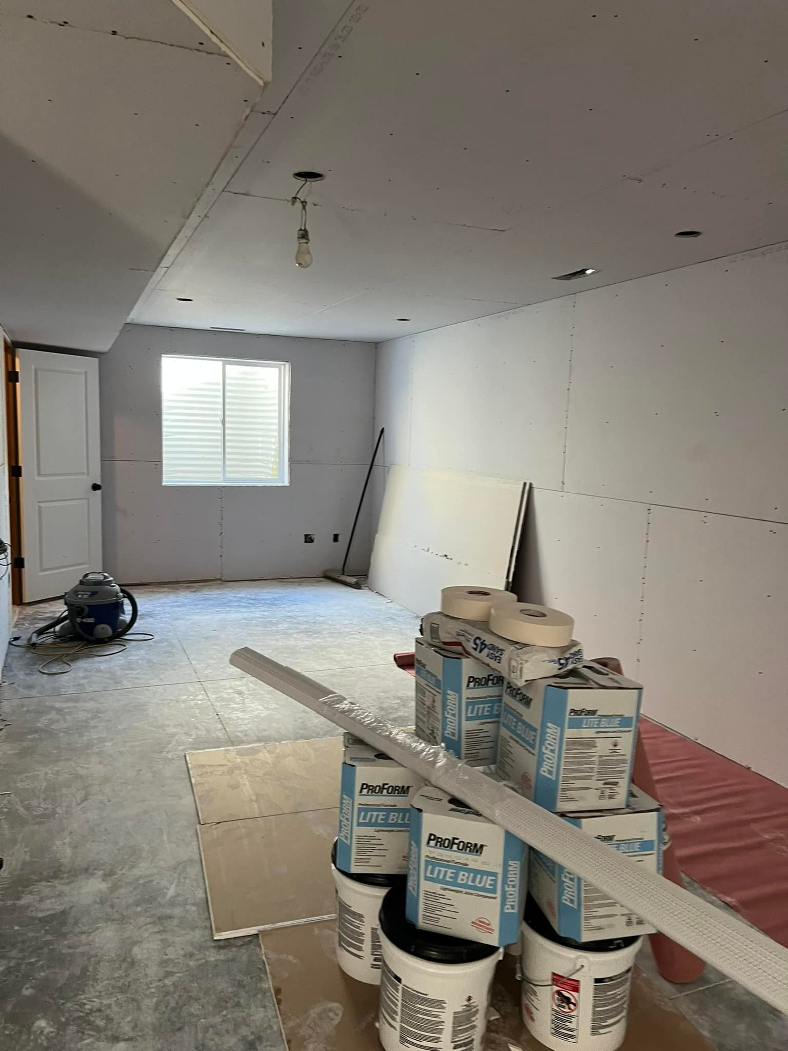 Room under renovation, drywall installed. Spackle buckets, tools, and a window are visible.