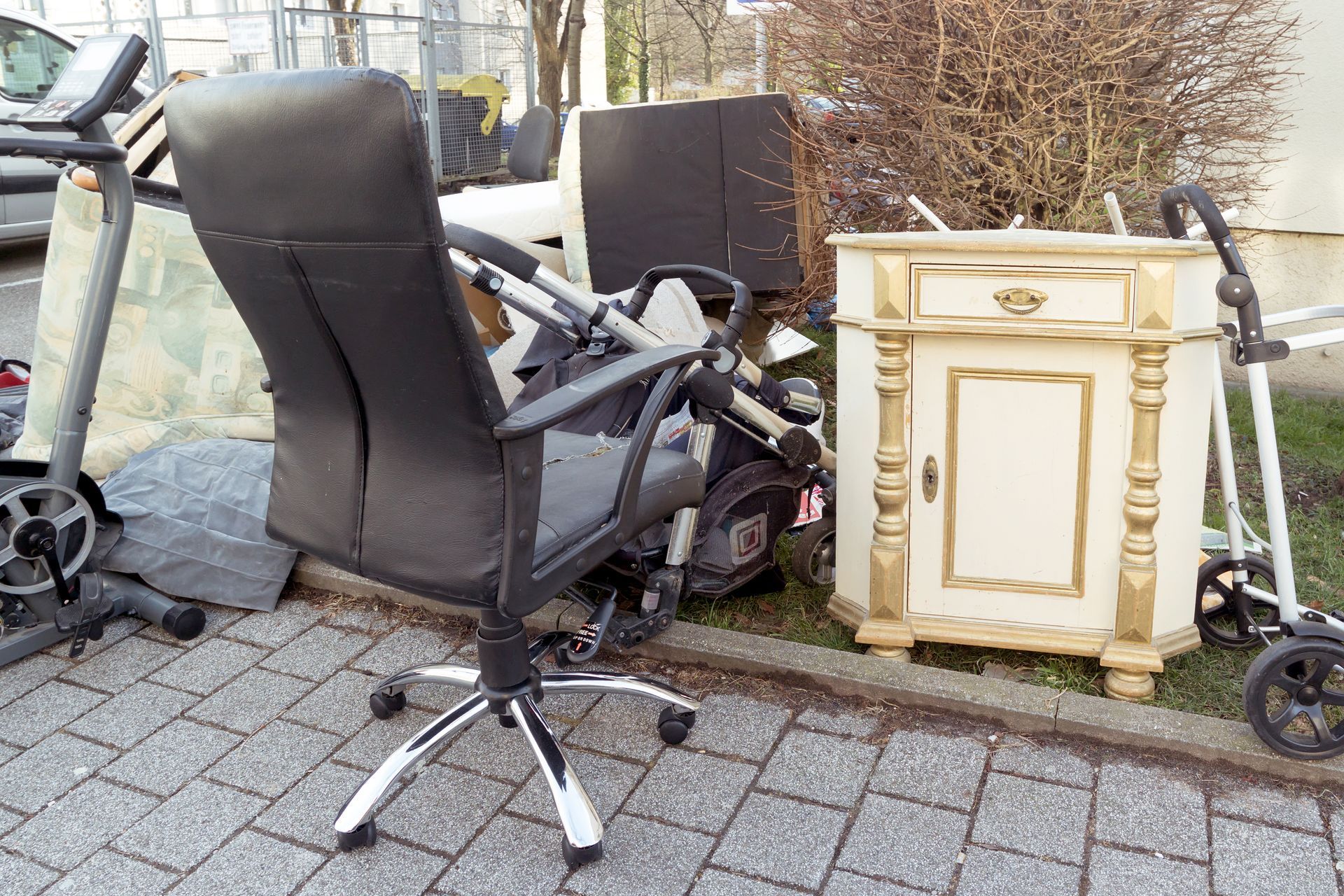 Discarded furniture and equipment, including a chair, cabinet, treadmill, and stroller, on a paved area near a curb.