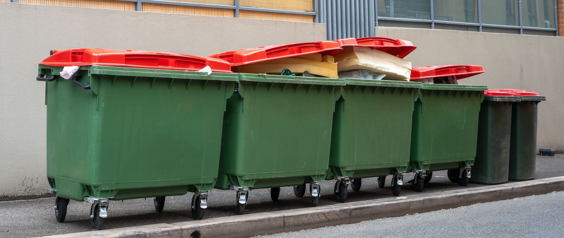 Several green dumpsters with red lids sit on a paved surface next to a building.