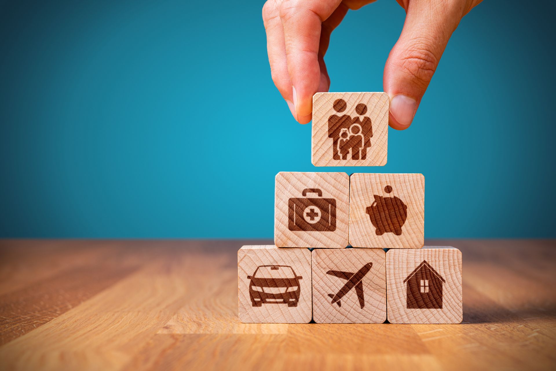Hand placing family icon block on top of insurance-themed blocks: car, house, plane, piggy bank, medical.