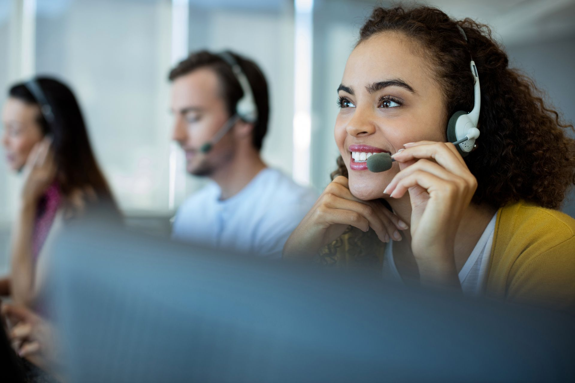 Woman with headset smiles while working at a desk, with colleagues in the background.