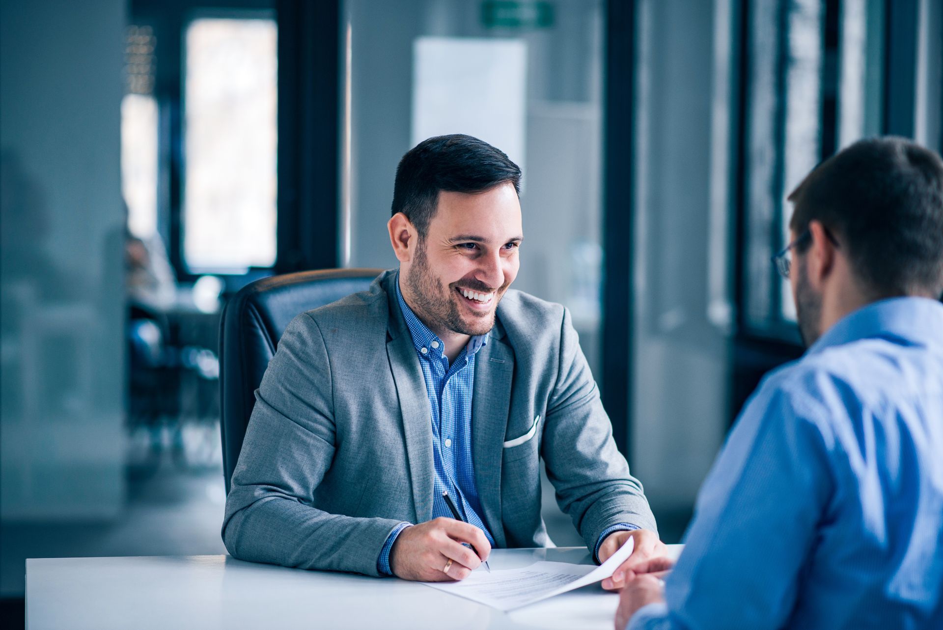 Man in suit laughs at documents with another person in an office setting.