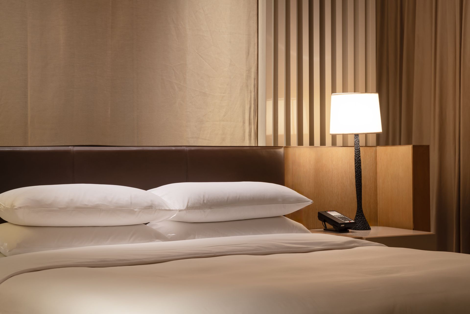 A neatly made bed with white linens and two pillows, next to a wooden nightstand with a lit lamp.