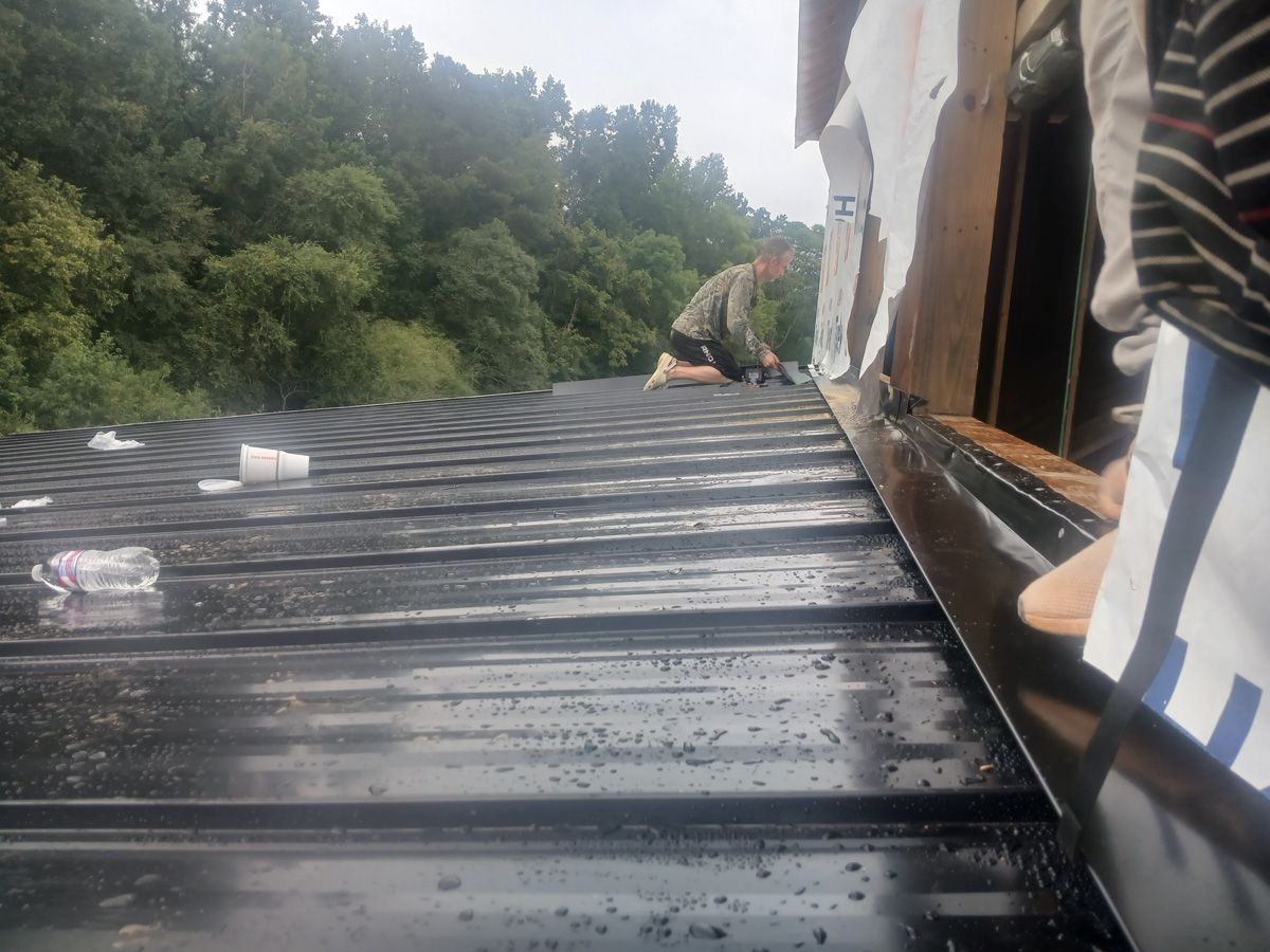 Person working on a black metal roof with a wooded background.