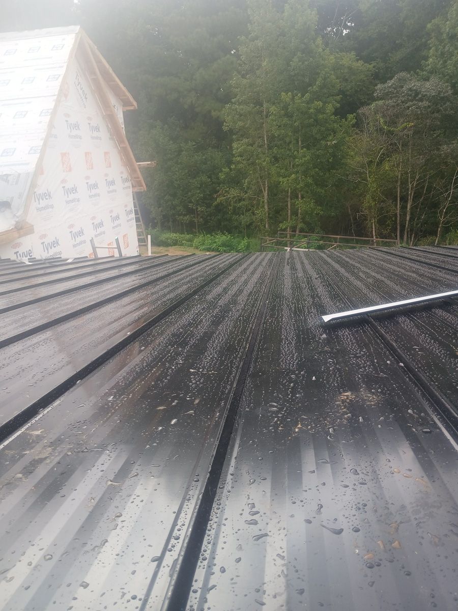 Black metal roof with water droplets, a house under construction, and a forested background.