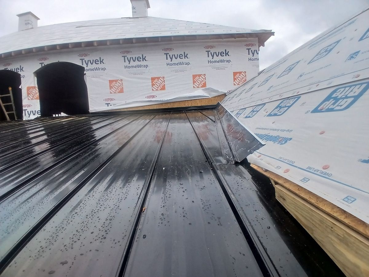 Metal roof in progress; Tyvek-covered walls. Black roof panels meet white underlayment. Cloudy sky.