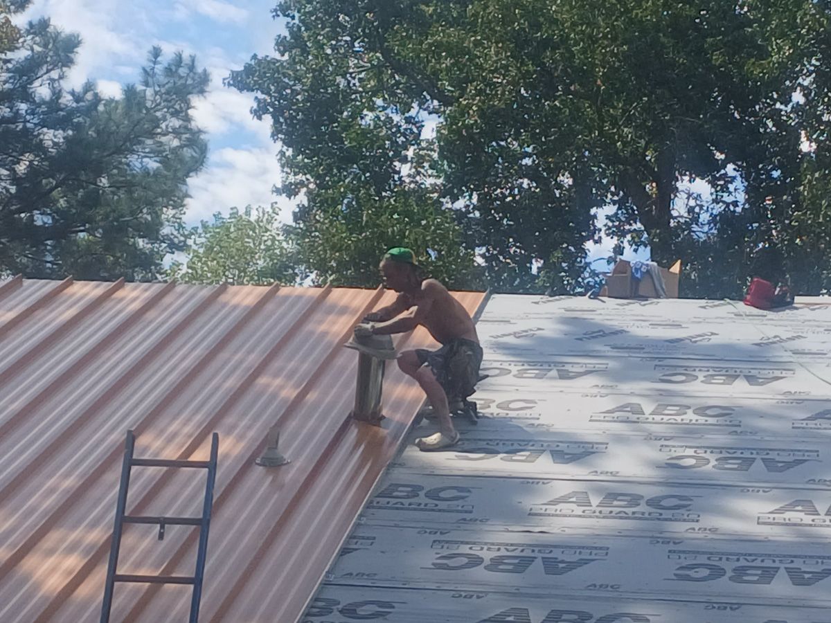 Man on roof installing copper metal roofing, near a ladder, under a blue sky.