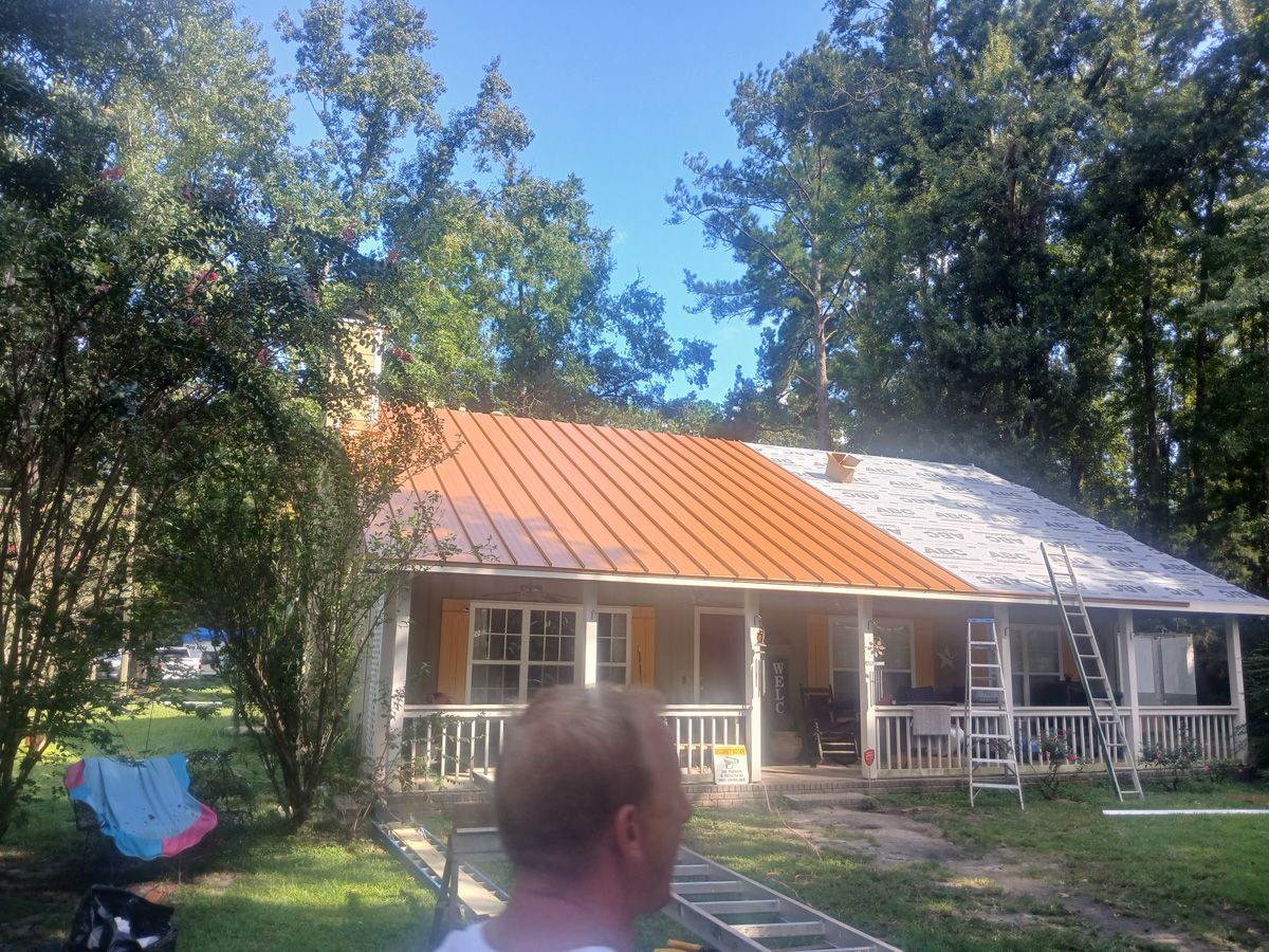 House with a partially replaced orange metal roof. Person in foreground. Trees surround.