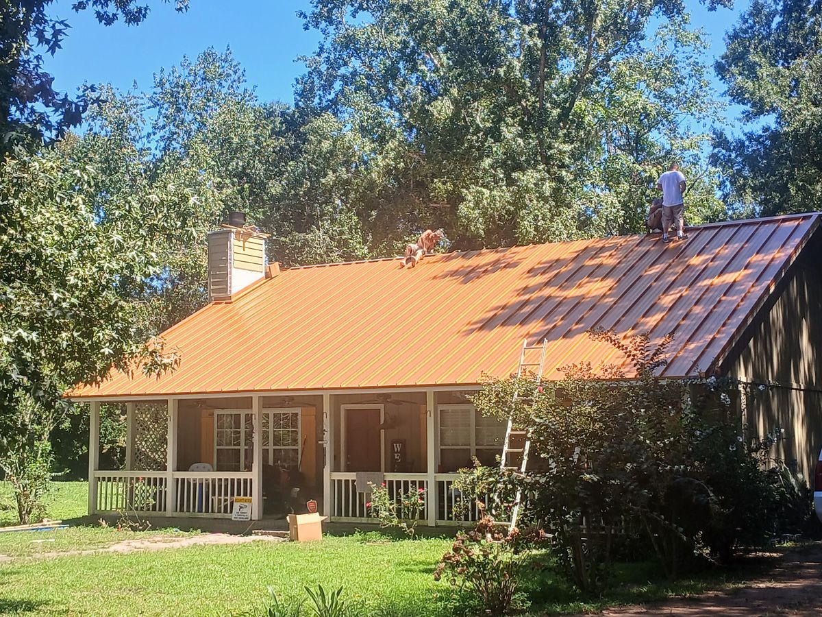 People on a roof installing orange roofing on a cabin with a porch; trees in the background.