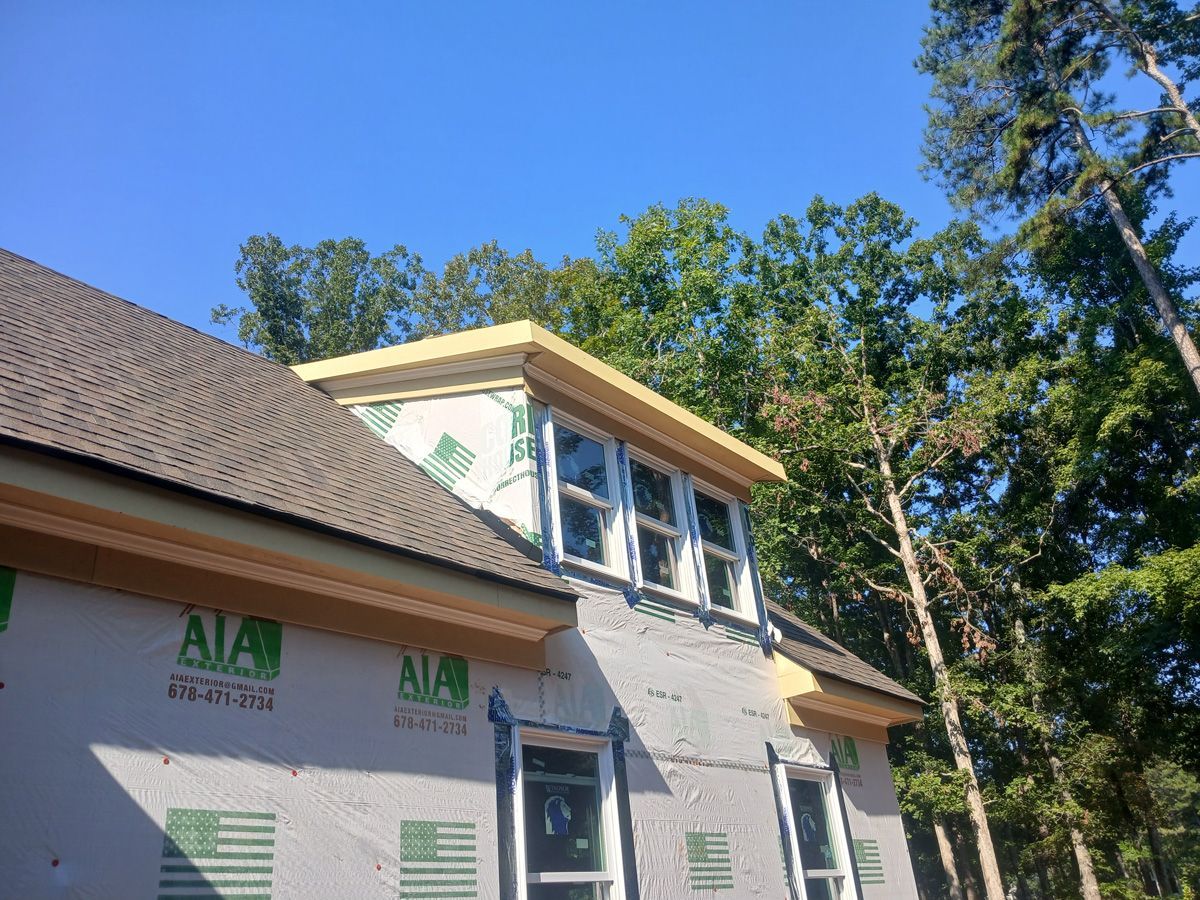 Construction of a house with a dormer; plywood roof and siding, windows, surrounded by trees under a blue sky.