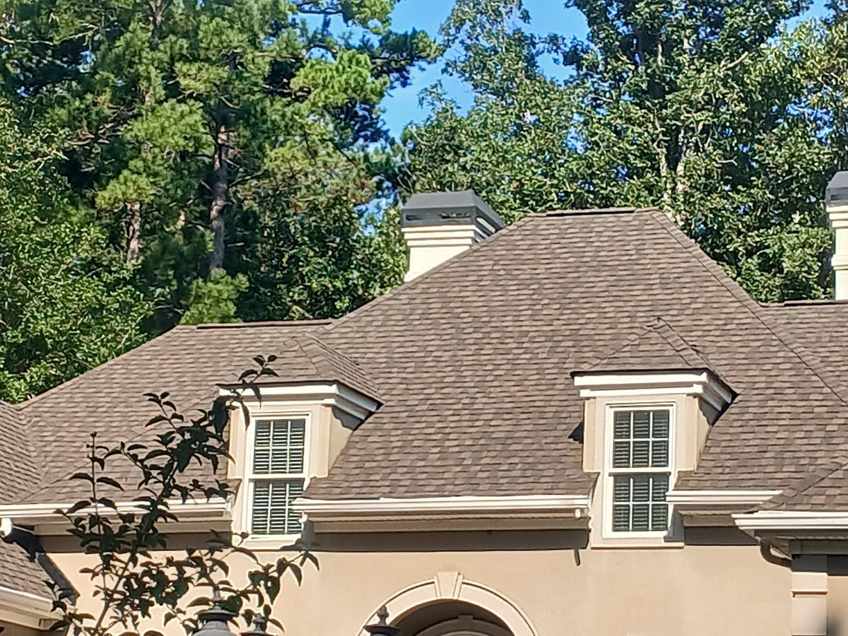 Brown shingled roof with dormers and chimney against a backdrop of green trees and a clear blue sky.