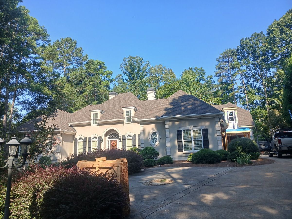 Beige house with a dark roof, surrounded by trees under a blue sky. A driveway leads to the house.