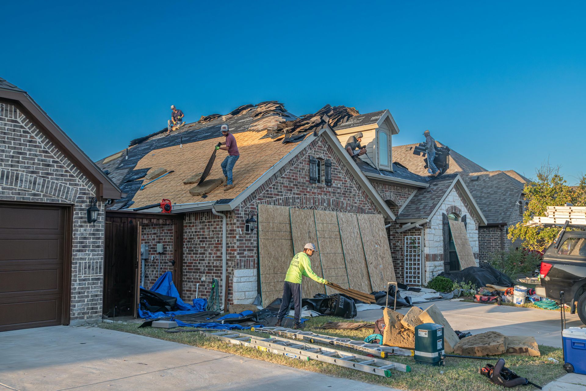 Roofers repairing a home with missing shingles; workers on the roof, plywood on the ground.