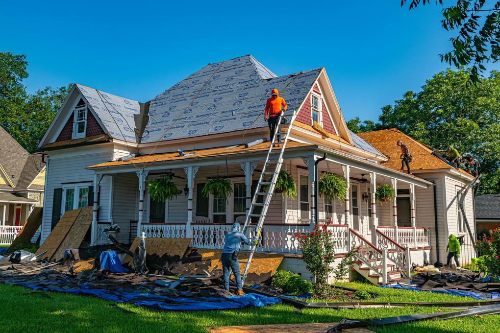 Roofers replacing shingles on a two-story white house with a porch on a sunny day.