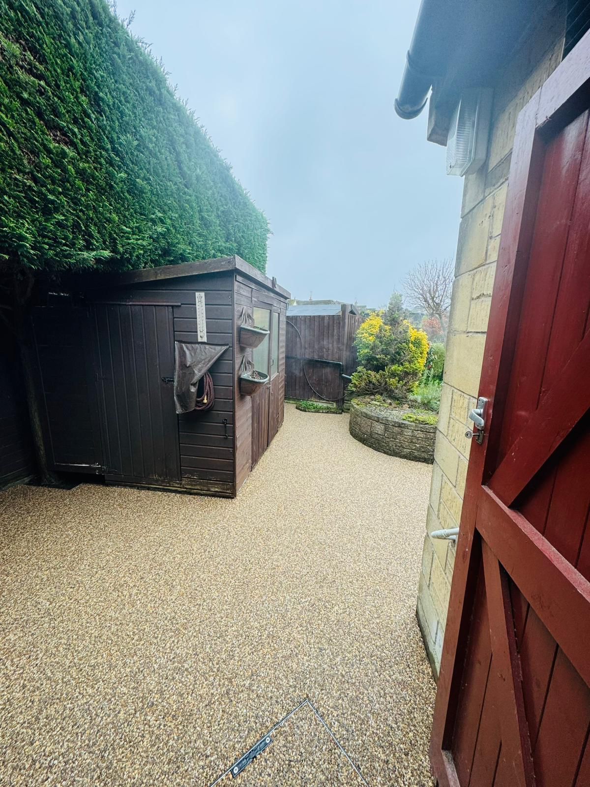 A gravel path leads between a dark wooden shed and a stone wall, framed by a red gate on the right.