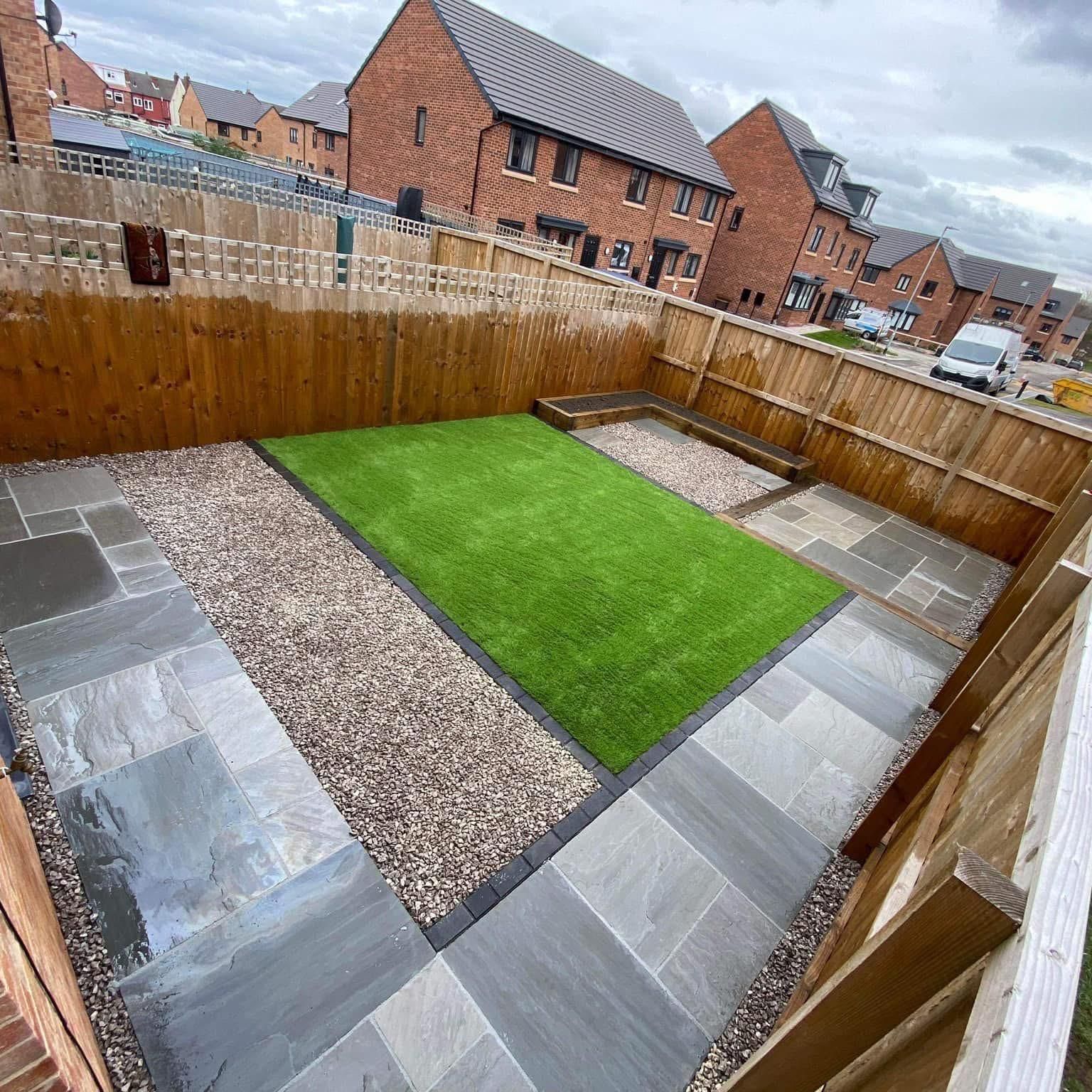 A small, modern backyard with gray paving stones, gravel, and an artificial grass rectangle. Wooden fence surrounds.