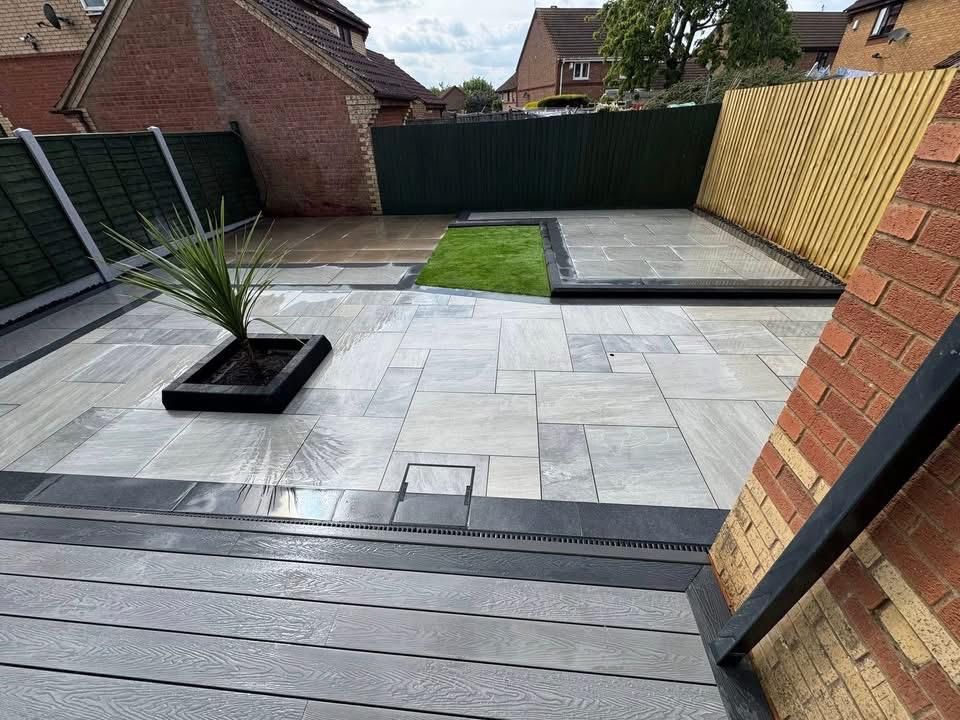 Patio with light gray paving stones, small grass patch, black planter, and brick wall.