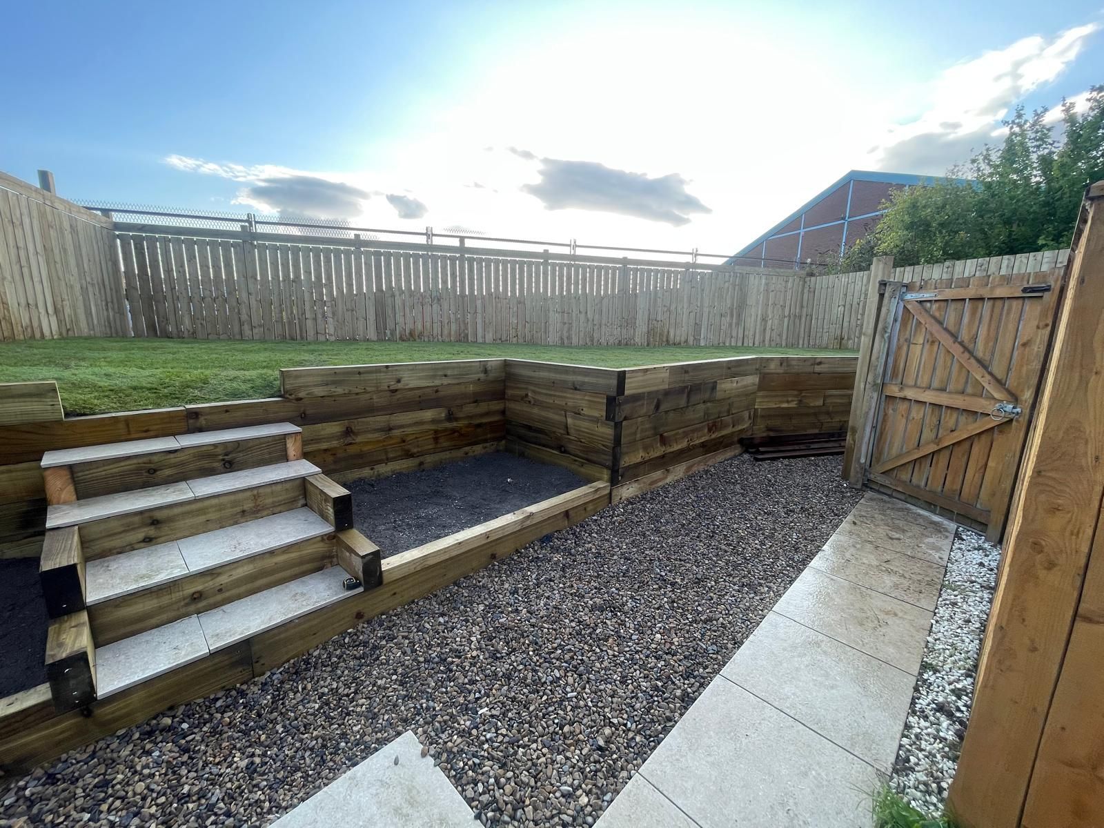 Wooden retaining walls and steps in a backyard with gravel and a gate leading to a wooden fence.
