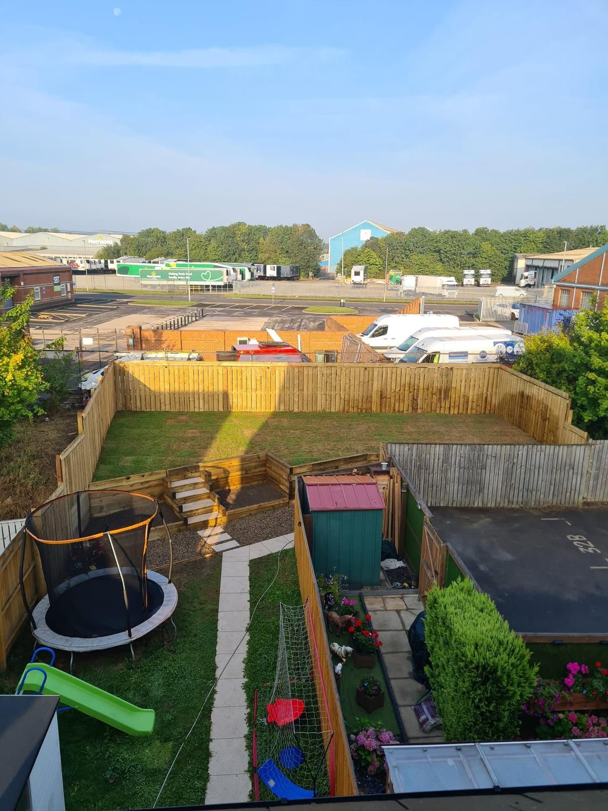 Backyard with wooden fences, green grass, a trampoline, slide, shed, and surrounding buildings under a blue sky.