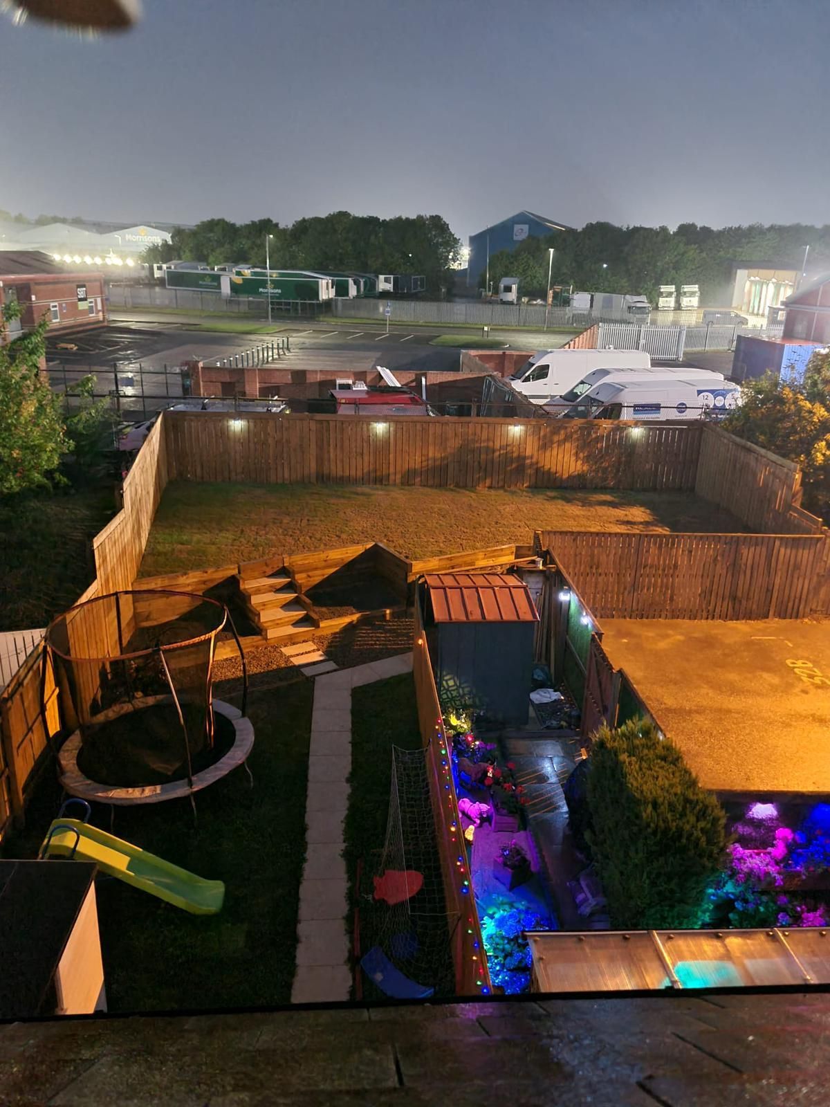 A backyard with a trampoline, slide, and lit fence, with buildings in the background at dusk.