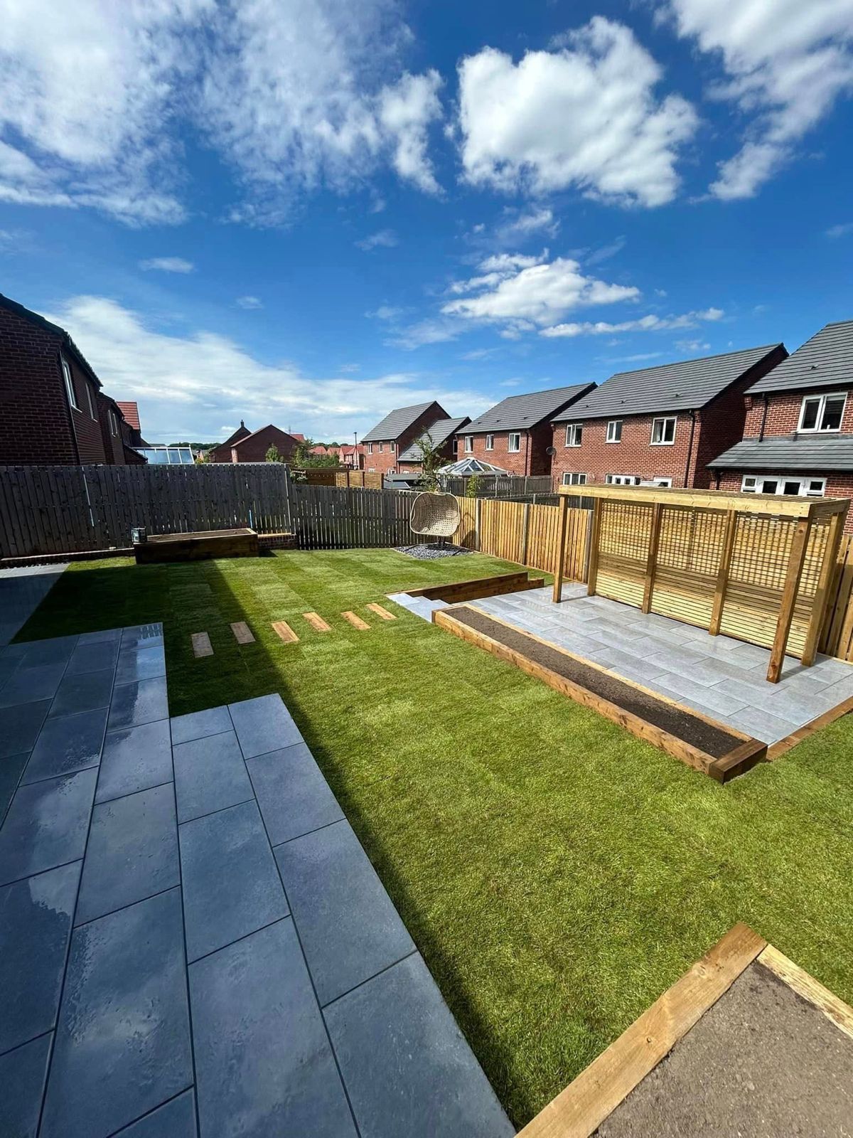 A residential backyard featuring a slate patio, a manicured lawn, a wooden trellis, and a clear blue sky.