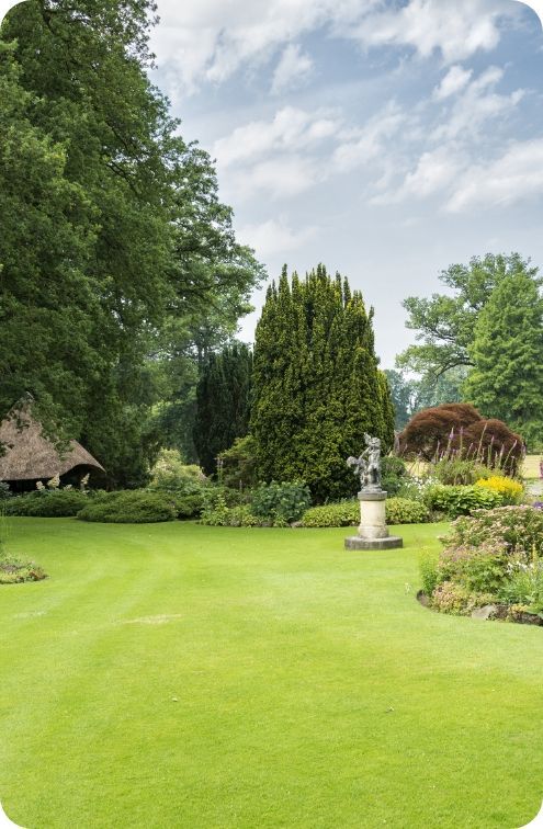 Lush green lawn in a garden with a statue, trees, and a building under a partly cloudy sky.