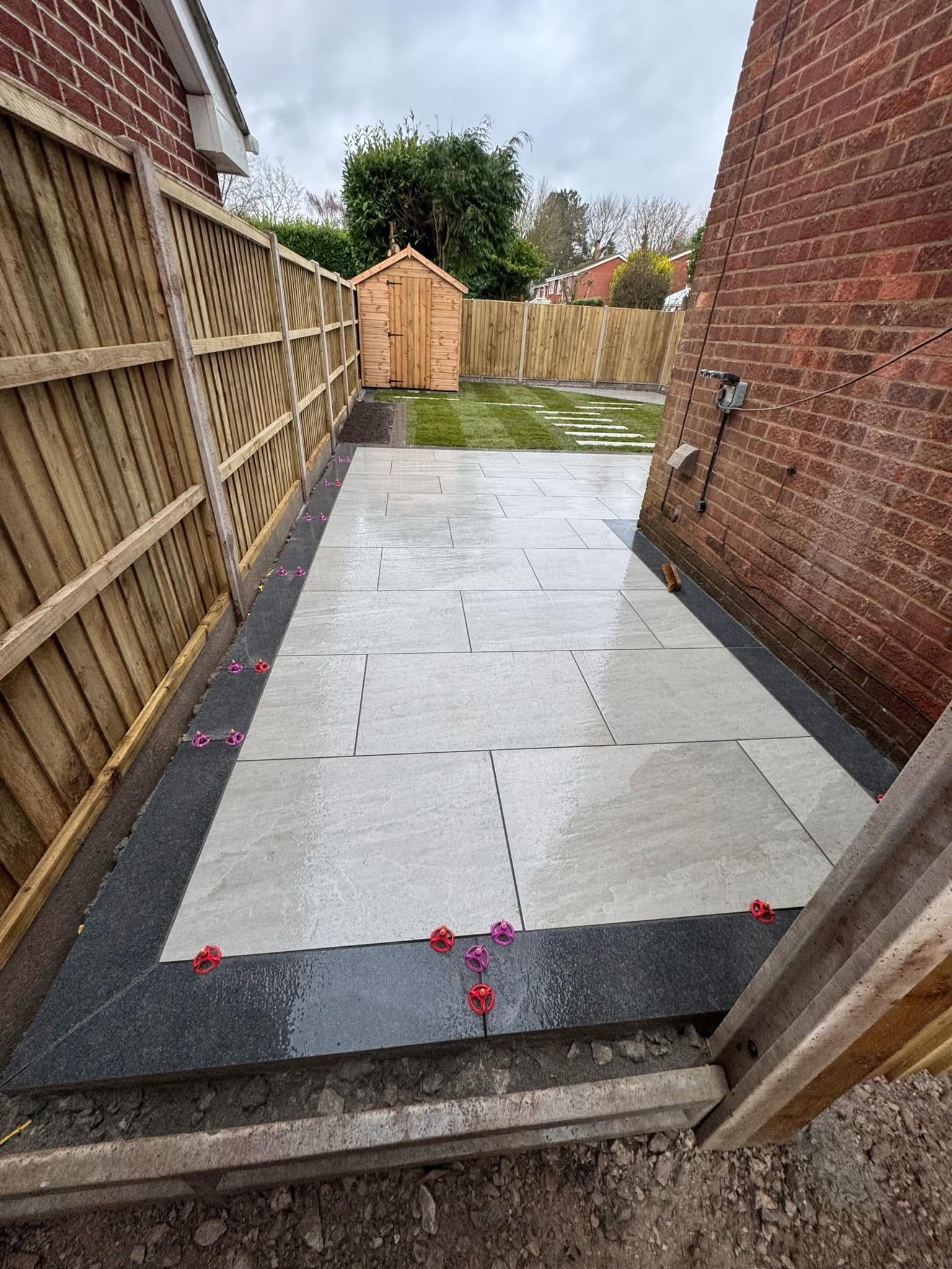 Newly paved backyard patio between a wooden fence and brick wall, with a shed in the distance.