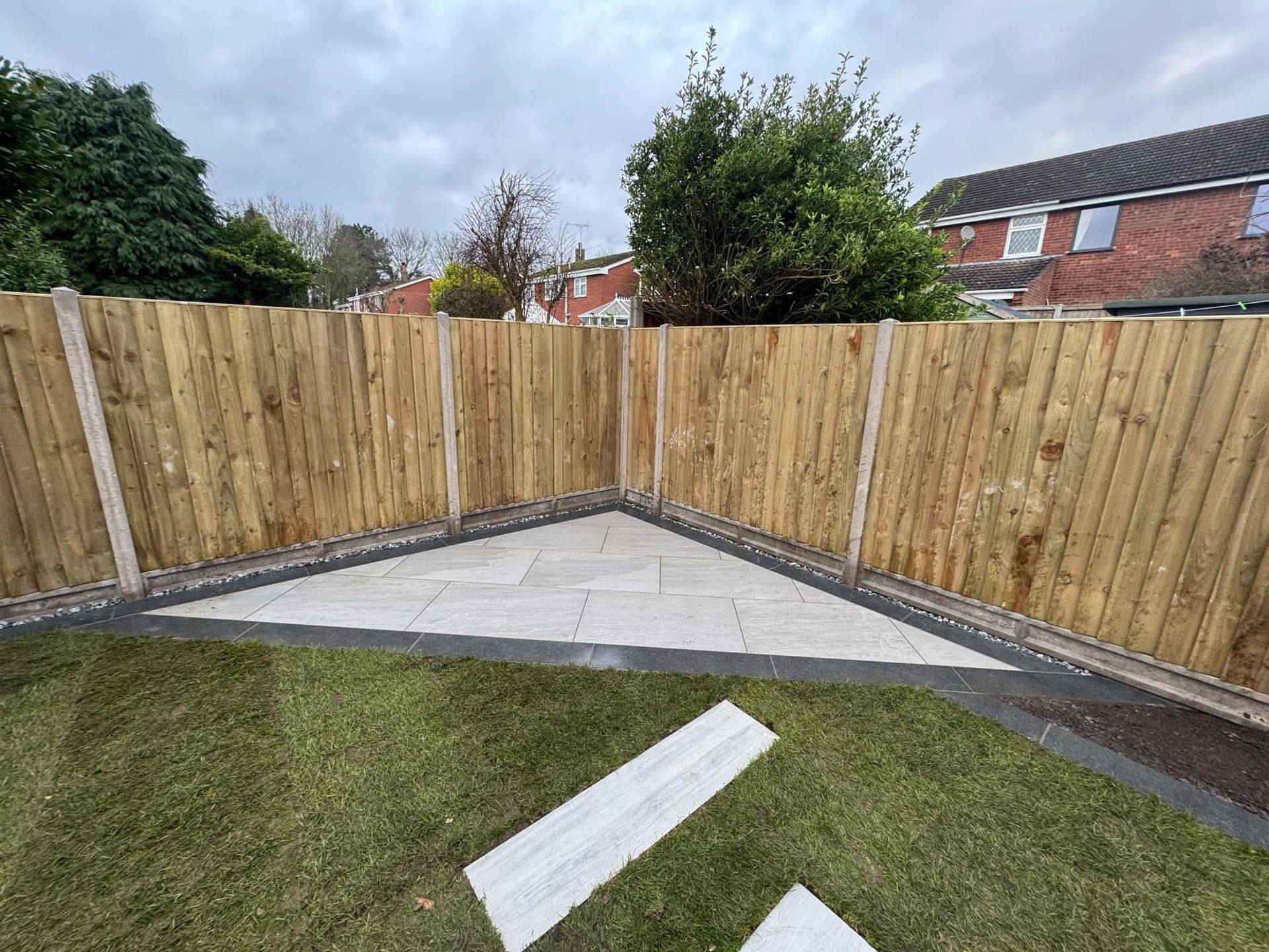 Wooden fence encloses a patio corner with gray pavers, surrounded by grass and houses.