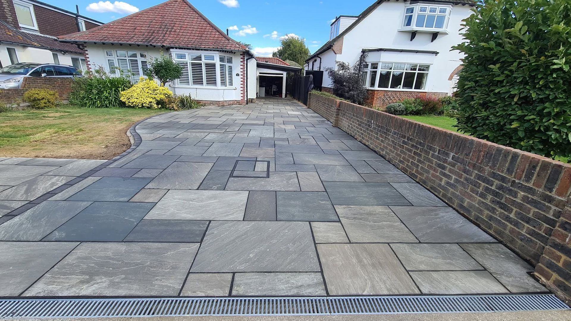 Driveway paved with gray rectangular stones, bordered by a brick wall and two houses.