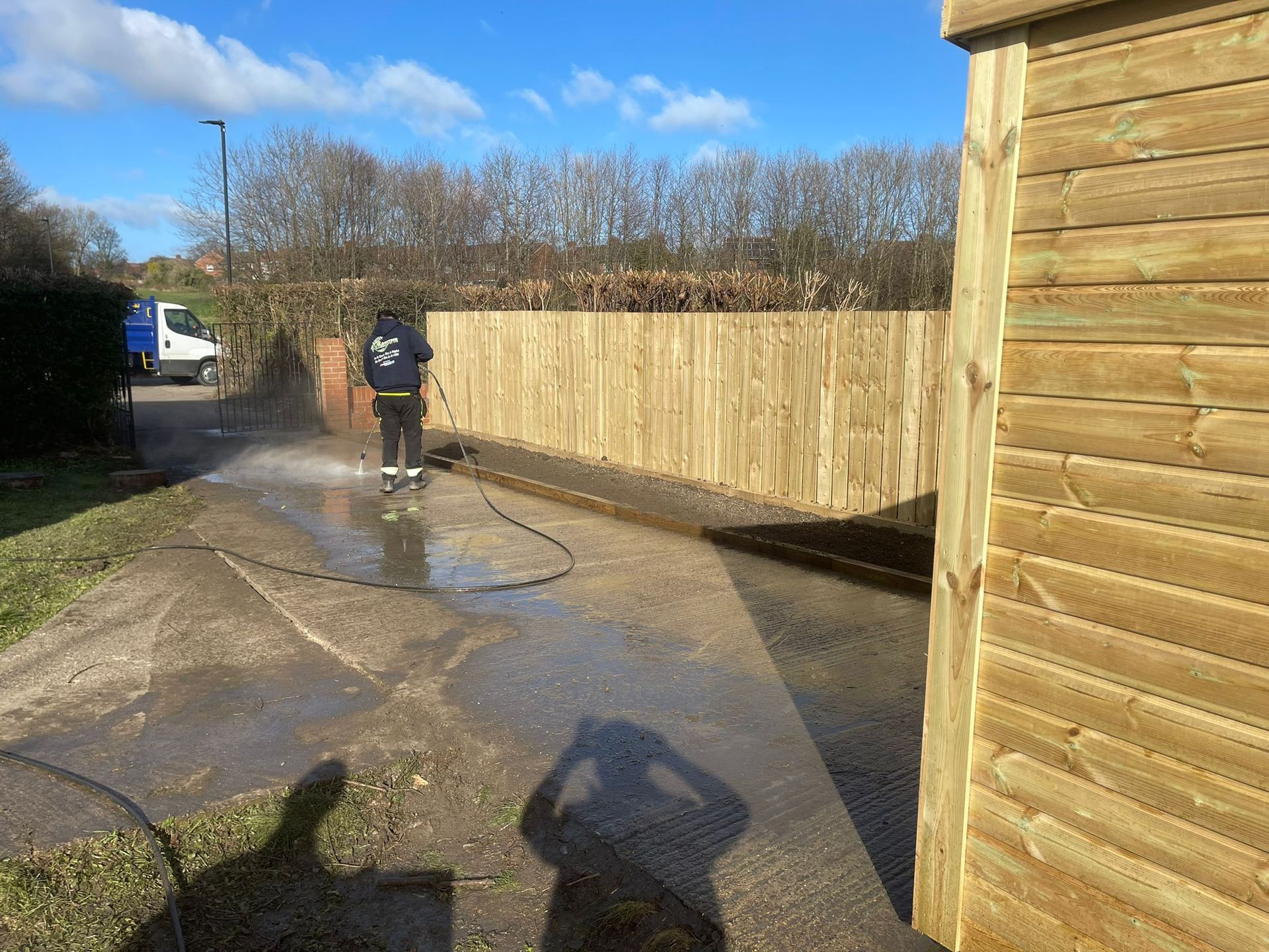 Person pressure washing a driveway next to a wooden fence on a sunny day.