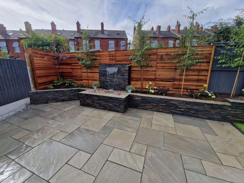 Patio with slate-faced planter, wooden fence, and water feature, with brick houses in the background.