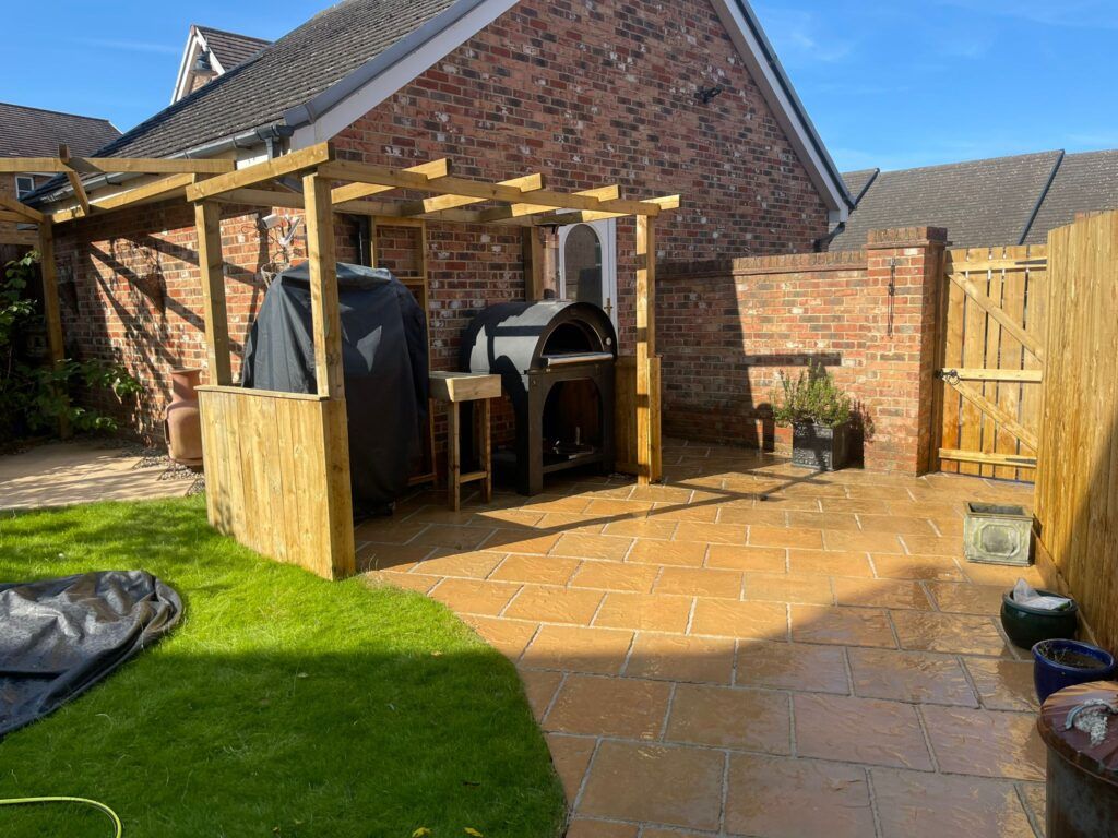 Outdoor pizza oven under a wooden pergola on a brick patio, surrounded by a lawn and brick building.