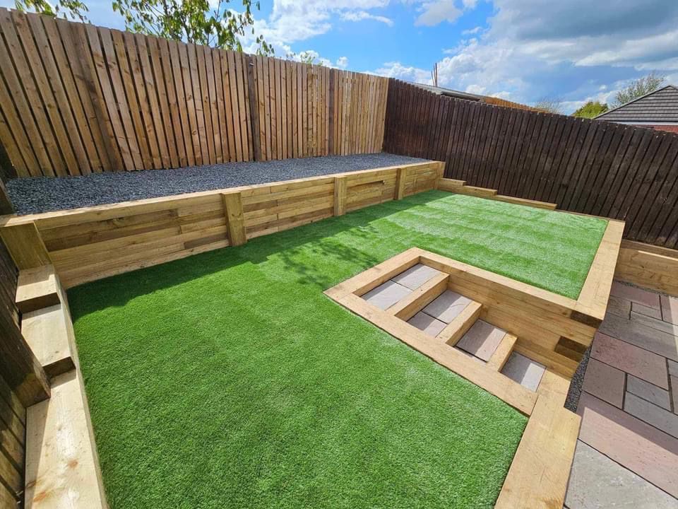 Lawn with retaining walls and steps built from wood. Brown fence and pavers in the background. Artificial grass.