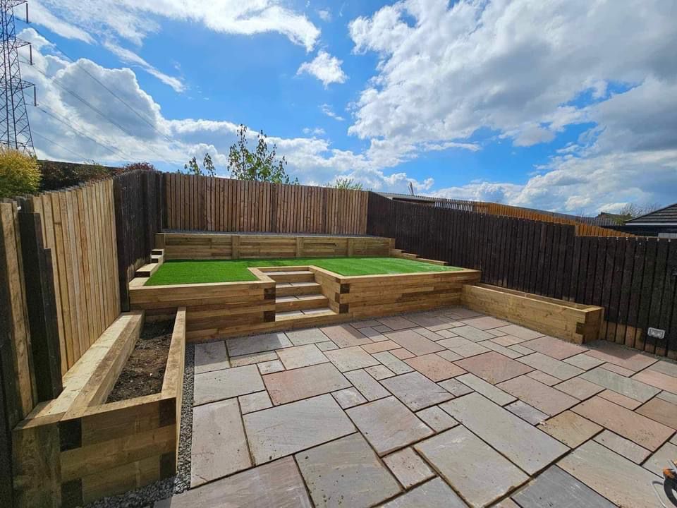 Patio with tiered garden beds, steps, and artificial turf, surrounded by wooden fences under a cloudy sky.