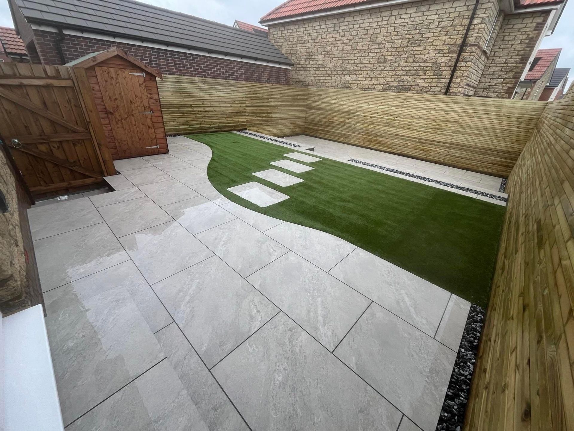 Backyard patio with gray paving, artificial grass strip, stepping stones, and wooden fence.