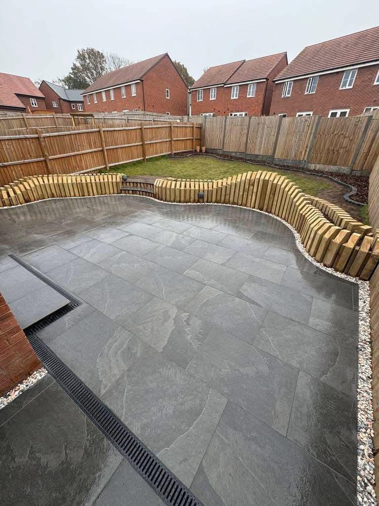 Patio with dark gray paving stones, wooden border, grass area, and houses in the background.
